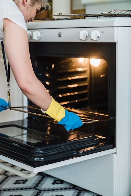 A person wearing a white shirt and yellow and blue cleaning gloves is cleaning the interior of a white oven with a wire rack inside. The oven door is open, revealing a clean, shiny black glass surface
